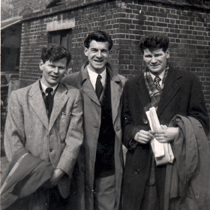 Three young men in overcoats standing outside building.