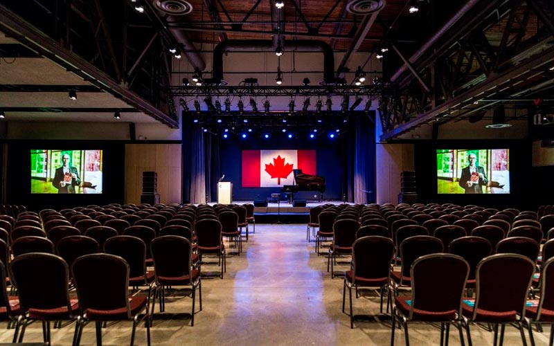 The venue space is set for a presentation and performance, with chairs theatre style and a centre aisle. On the stage there is a podium, a black grand piano, and a Canadian flag backdrop. Two projected screens are set on both sides of the stage.