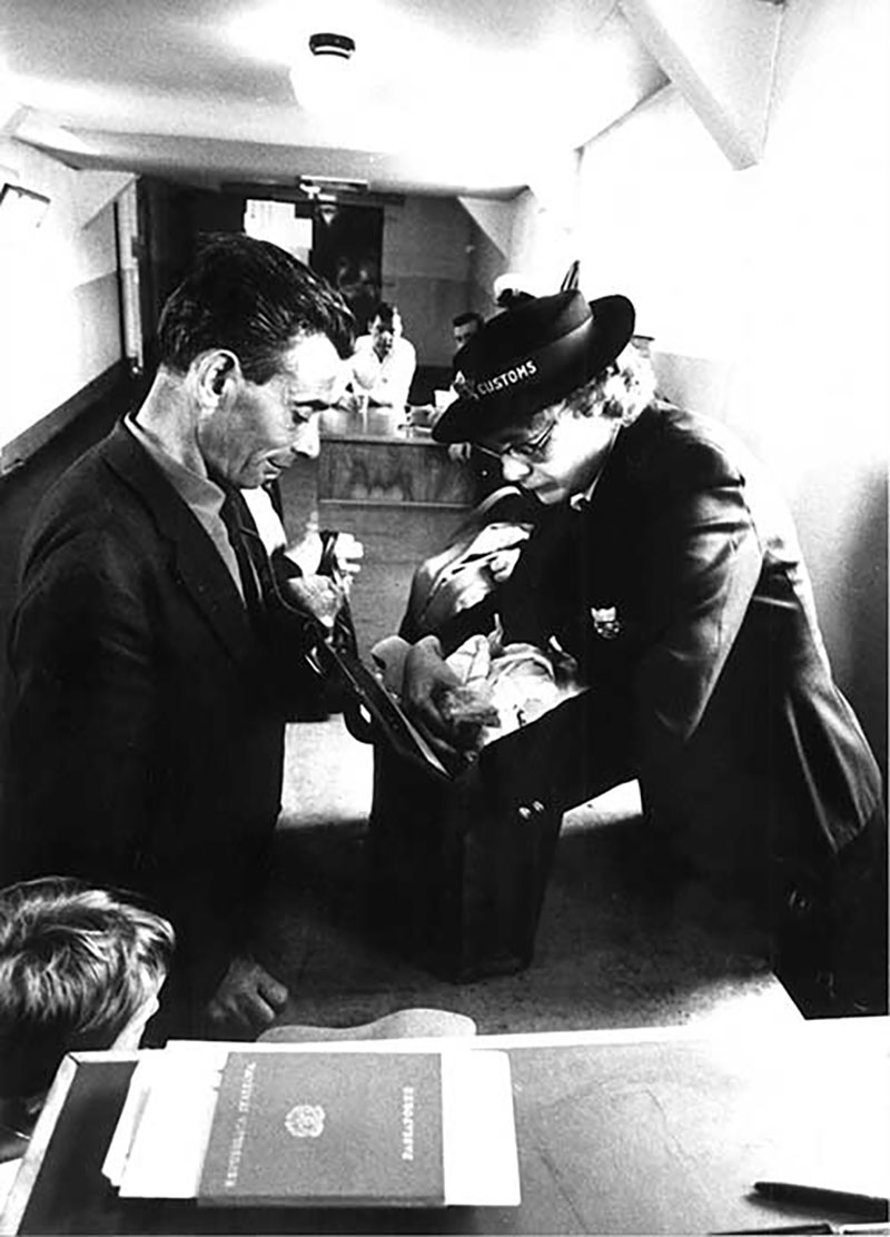 A female customs officer searches though the baggage as the owner of the baggage looks on.