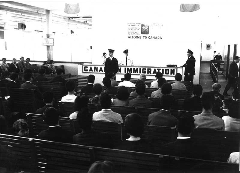 Two officers stand in front of an immigration desk and there are several people seated on benches facing them.