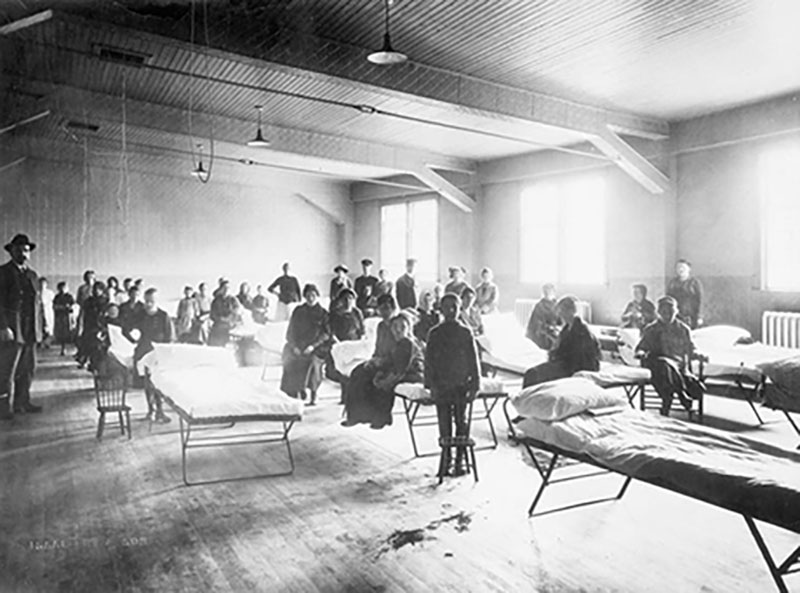 A large dormitory is filled with makeshift beds and people sitting on them.