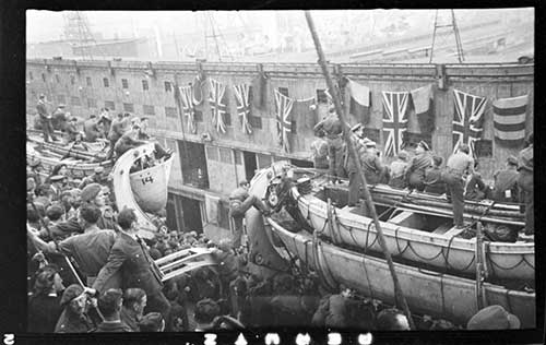 A ship filled with men in uniform docking next to a building strung with Union Jack flags.
