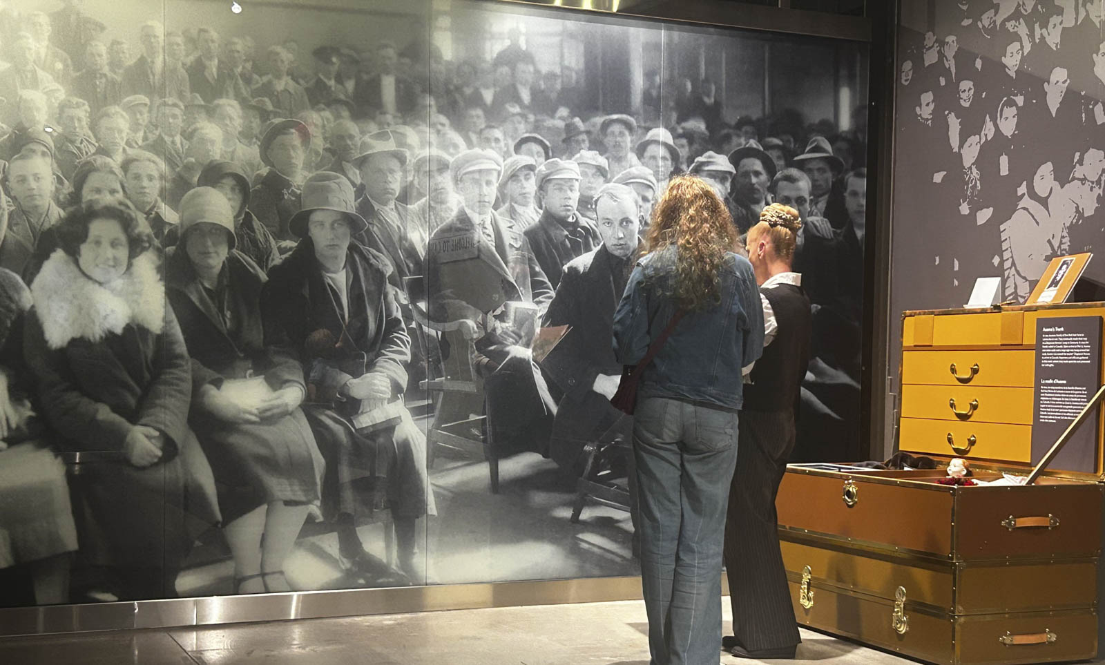 Two women with their backs to us stand in front of a wall-sized historical image of a large crowd waiting on benches.