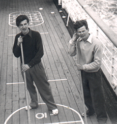 Two men standing are playing shuffleboard while on the ship's deck.