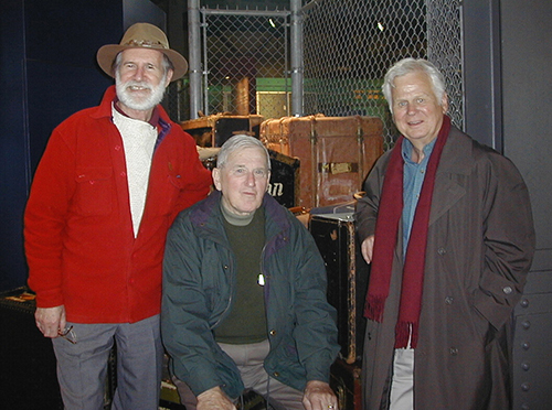 Three older men wearing jackets stand together.
