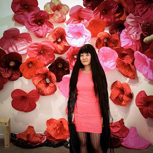 Beautiful big flowers made of fabric cover a wall. A woman with very long dark hair stands in front of the wall.