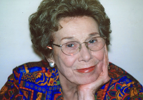 An elderly woman wearing glasses rests her chin on her hand and smiles at the camera.