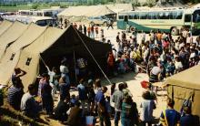 Large groups of Kosovar refugees waiting outside of military tents, with busses in the background.