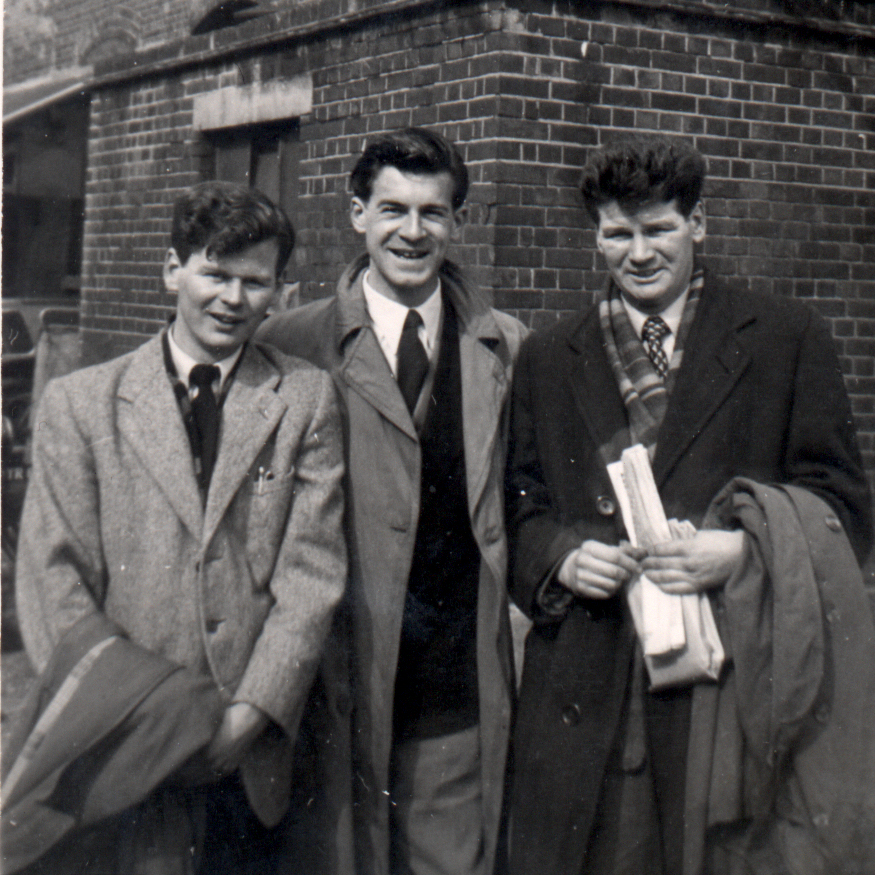 Three young men in suits and overcoats, standing in front of a building.