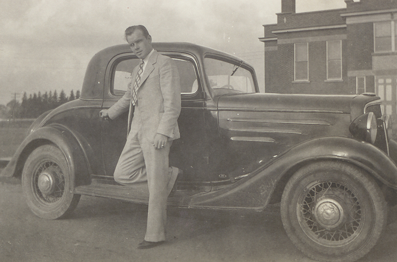 A man in a suit and tie stands against a car, holding the door handle.