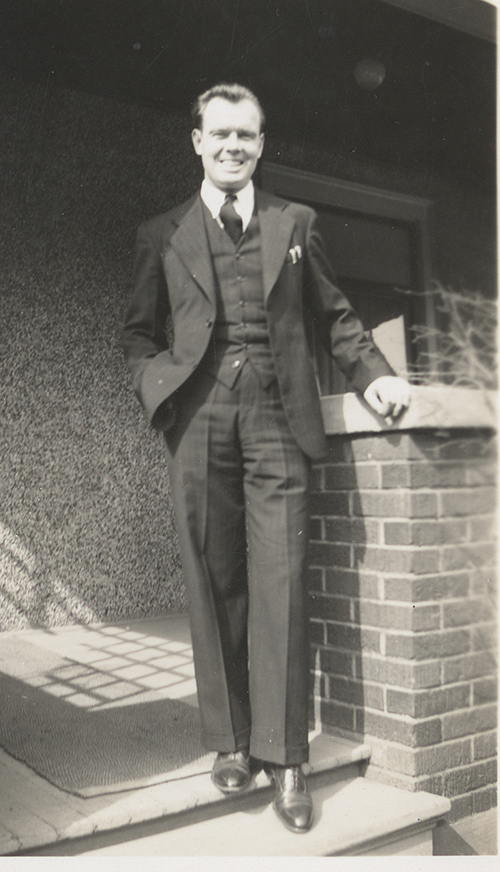 A smiling man in a three-piece suit stands on the steps of a building.