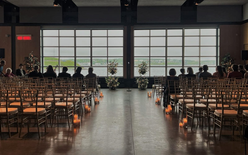 White chairs are set in a theatre style for a wedding ceremony facing windows that showcase a view of the harbour. Two floral stands and a mic stand are at the end of aisle. Candles in cylinder vases are placed next to chairs down the aisle.