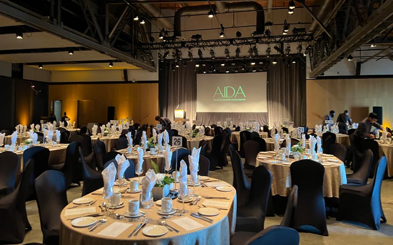 A venue with tall ceilings is set for a banquet with oval tables and banquet chairs. On the stage there is a podium and projection screen, and stage lights are focused on the podium. 
