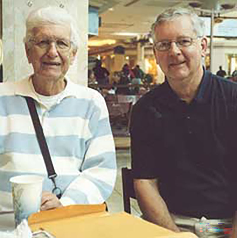Two bespectacled men are seated at a table, both smiling directly at the camera.