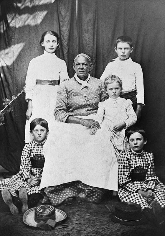 An older Black woman wearing an apron is surrounded by five white children. One child rests on her lap.