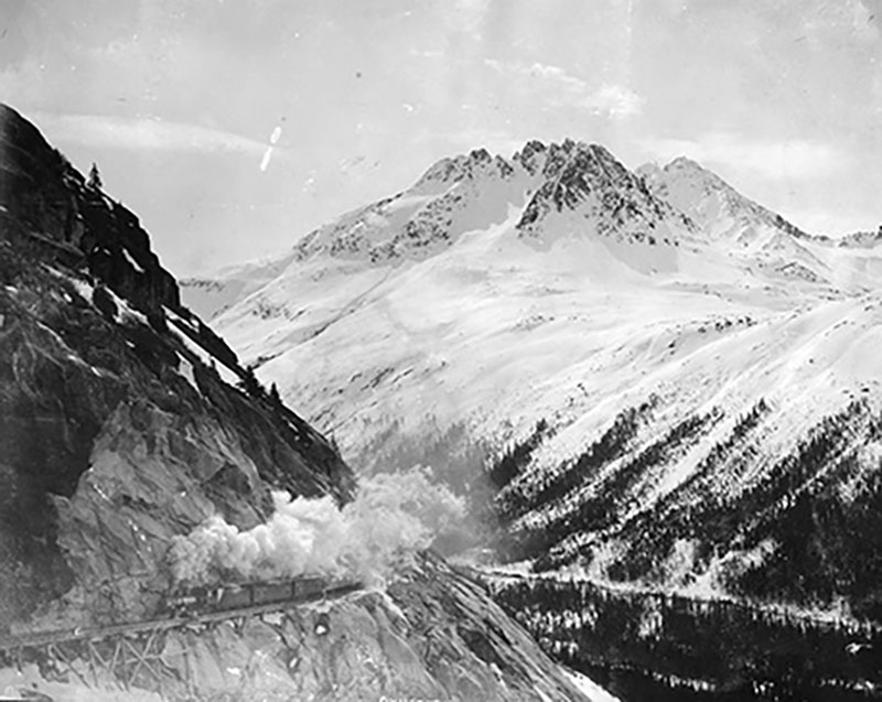 A train with smoke billowing out is slowly moving along a mountain pass.