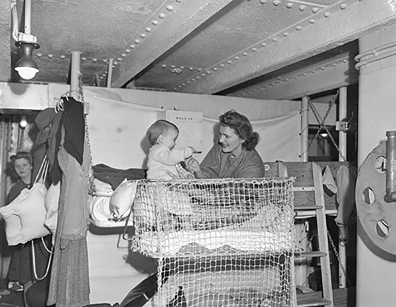 A woman sits on a bed in a ship's berth and plays with her baby.