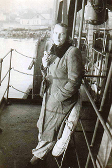 A lady wearing a long winter coat stands on a ship's deck.