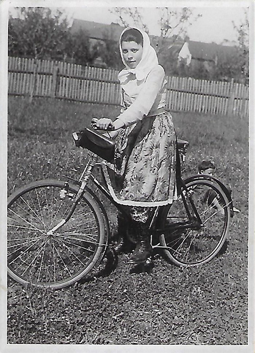 Black and white photograph of a young girl on a bike. She is dressed in a long-sleeved skirt and sweater and wears a scarf on her head.